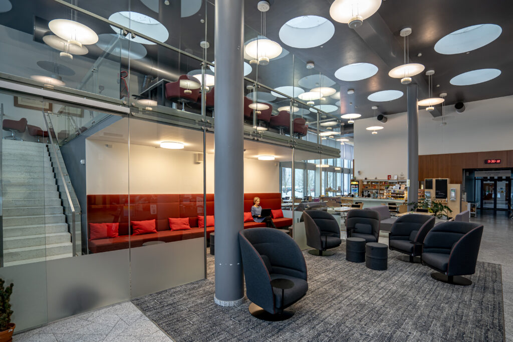 Avatud puhkeala hallide lounge-toolide, punase istumisnurgaga ja klaasvaheseintega; taustal kohvikuala
