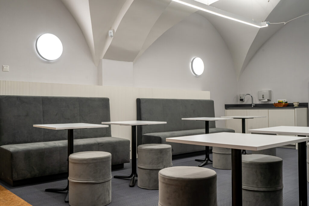 Valge võlvlaega puhkeruum hallide diivanite, valgete laudade ja ümarate tumbadega; taustal väike köögiala valamu ja tarvikutega